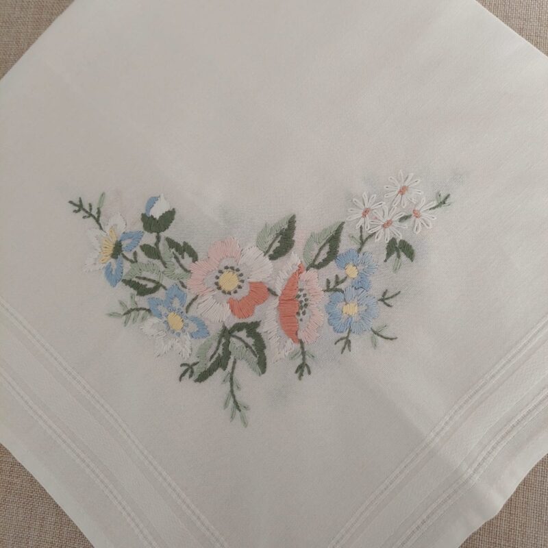 Vista de primer plano de una esquina de la carpeta de mesa de algodón cuadrada blanca, mostrando en detalle el ramillete de flores silvestres bordado a mano en tonos rosa, celeste y durazno, junto a una guarda calada lineal.