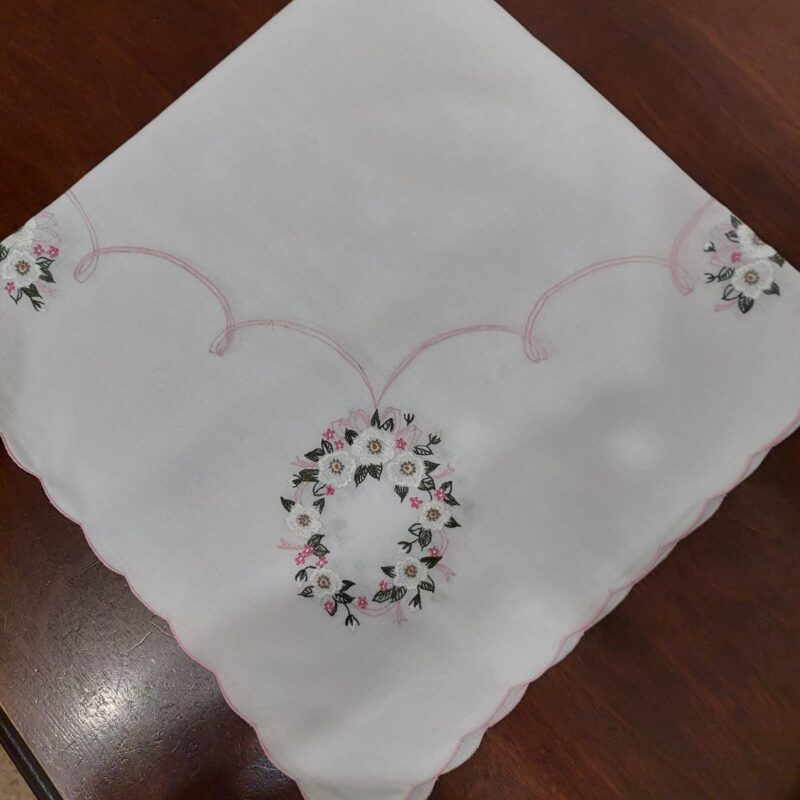 Vista de una carpeta de mesa cuadrada blanca sobre una mesa de madera oscura, mostrando su diseño esquinado de guirnaldas de margaritas bordadas y borde festoneado rosa con un florero encima.