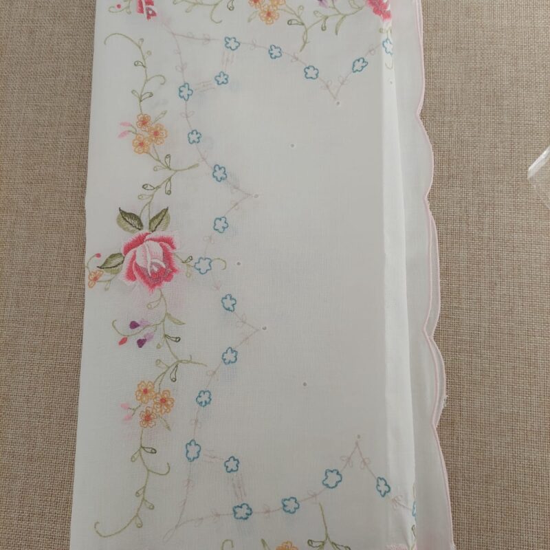 Vista detallada de una carpeta de mesa cuadrada blanca extendida, mostrando su profuso diseño de rosas fucsias bordadas a mano, guías florales multicolores y un campo central decorado con pequeñas flores celestes.