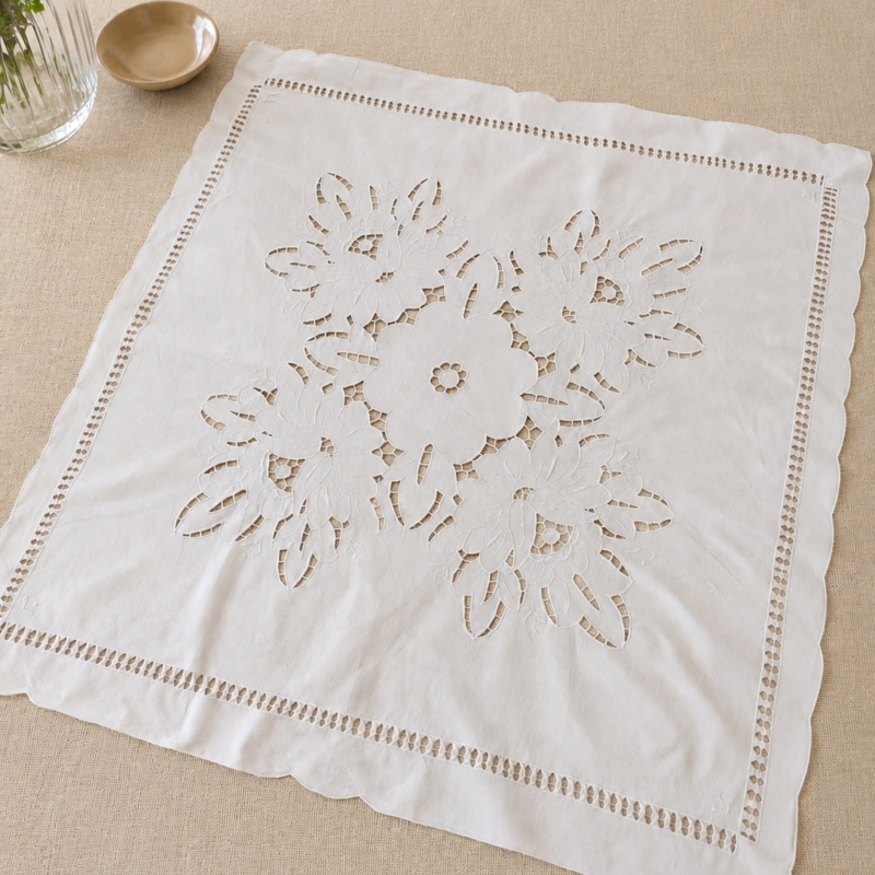 Vista general de una carpeta de mesa cuadrada blanca extendida, que muestra un intrincado y gran diseño central calado a mano estilo Richelieu con motivos de flores simétricas, enmarcado por una línea de vainicas y bordes ondulados festoneados.