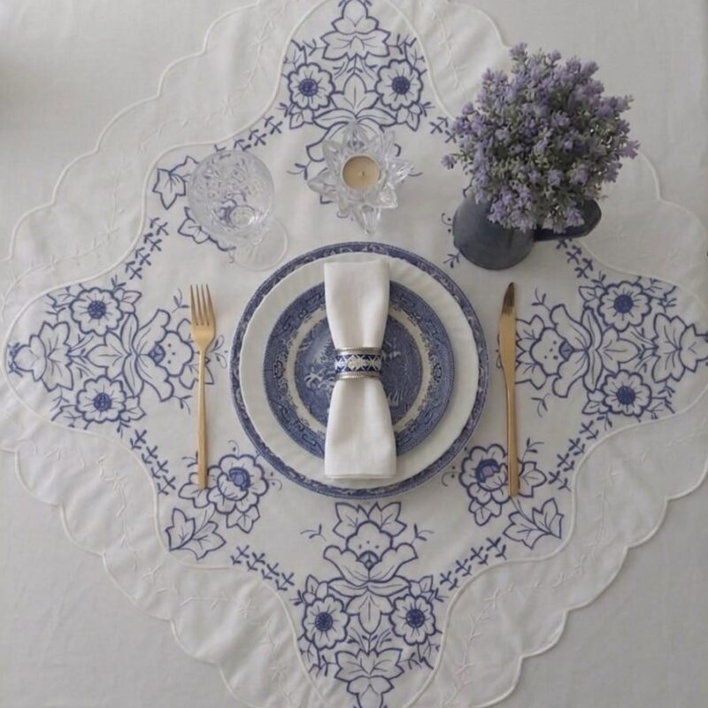 Vista de primer plano de una carpeta de mesa cuadrada blanca doblada, mostrando en detalle su profuso bordado floral en hilo azul cobalto, pequeñas guías caladas en cruz y un borde festoneado con bordado blanco.