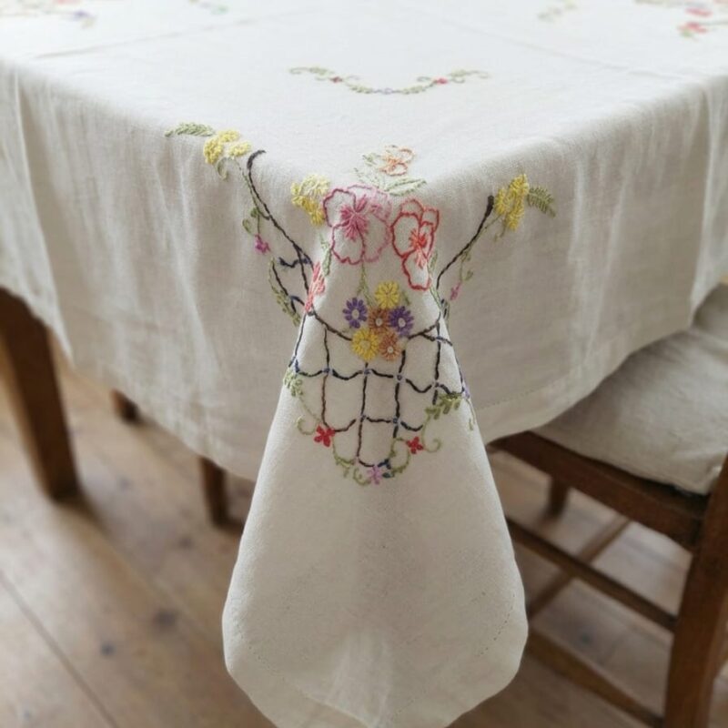 Vista general de un mantel rectangular blanco extendido, mostrando sus cuatro esquinas decoradas con un patrón de enrejado negro y frondosos ramilletes de flores bordadas a mano en tonos fucsia, amarillo, azul y violeta.
