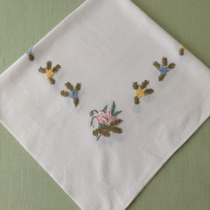 Vista de una carpeta de mesa de hilo blanco sobre un mantel verde, mostrando su diseño esquinado de flores bordadas a mano y detalles decorativos con un centro de mesa encima.