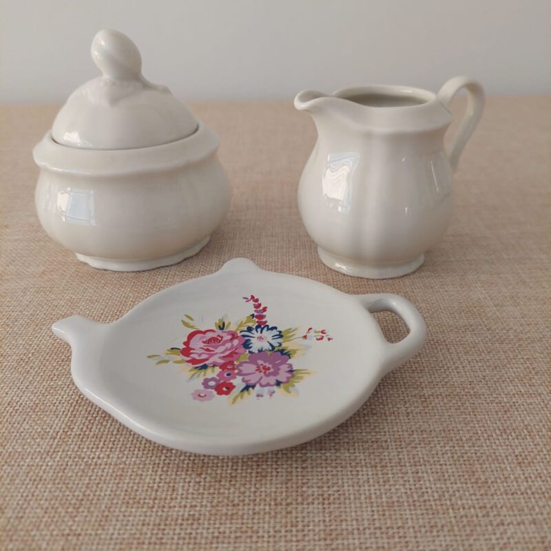 Mini set de servicio de té vintage sobre mantel que incluye un azucarero con tapa y un lechero de porcelana blanca alemana Seltmann Weiden, junto a un platillo posa bolsas de té Price & Kensington con forma de tetera e ilustración de rosas.