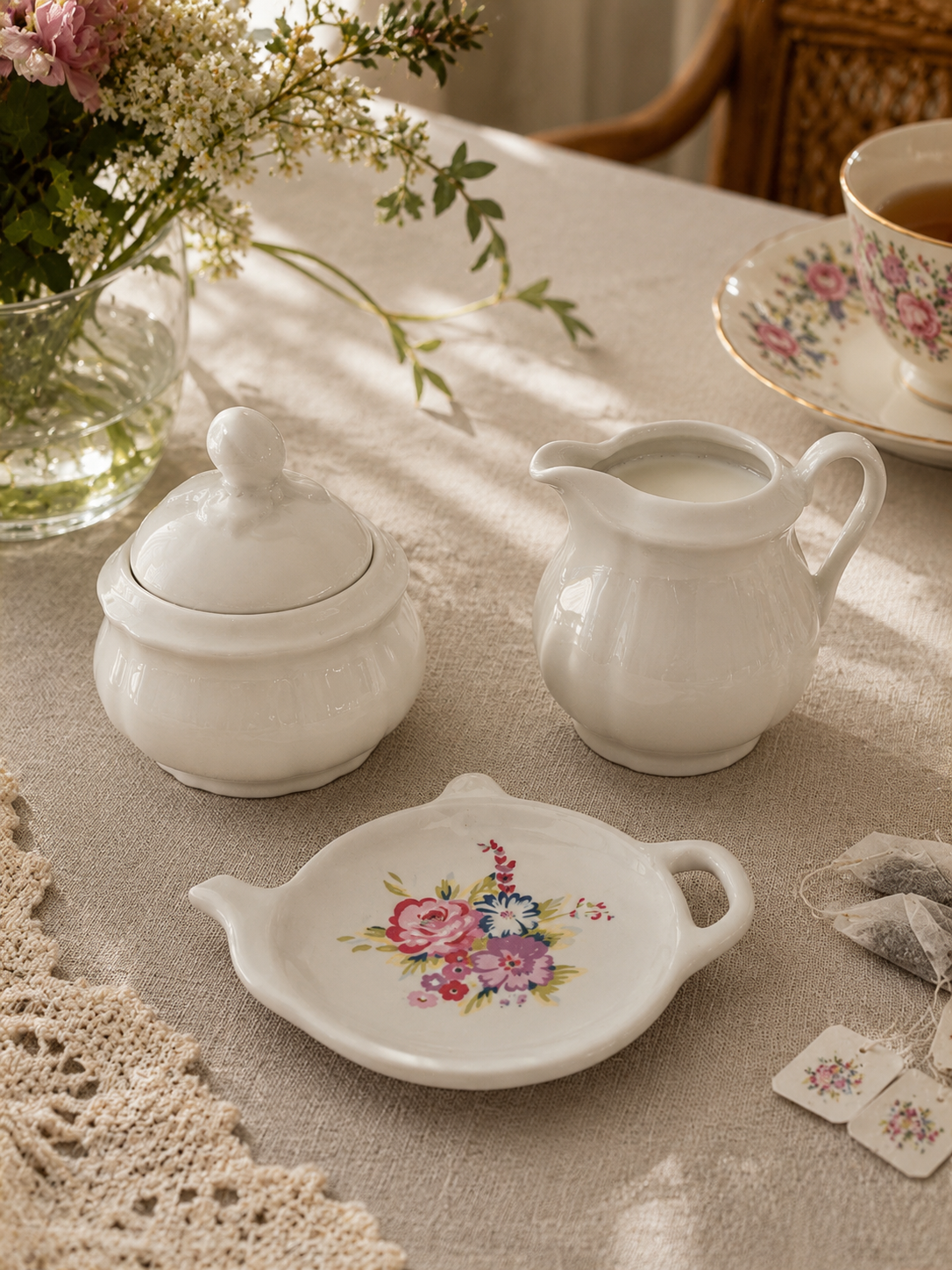 Mini set de servicio de té vintage sobre mantel que incluye un azucarero con tapa y un lechero de porcelana blanca alemana Seltmann Weiden, junto a un platillo posa bolsas de té Price & Kensington con forma de tetera e ilustración de rosas.
