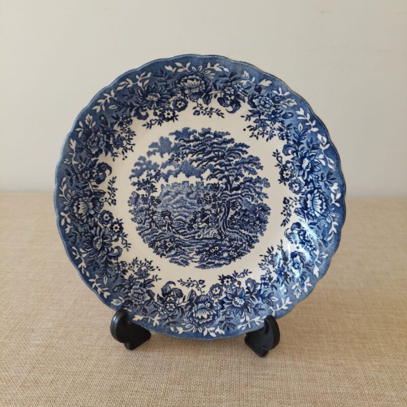 Vista frontal de un plato hondo festoneado de loza inglesa Alfred Meakin con diseño azul y blanco del patrón "The Courtship", mostrando una escena campestre central y bordes florales.