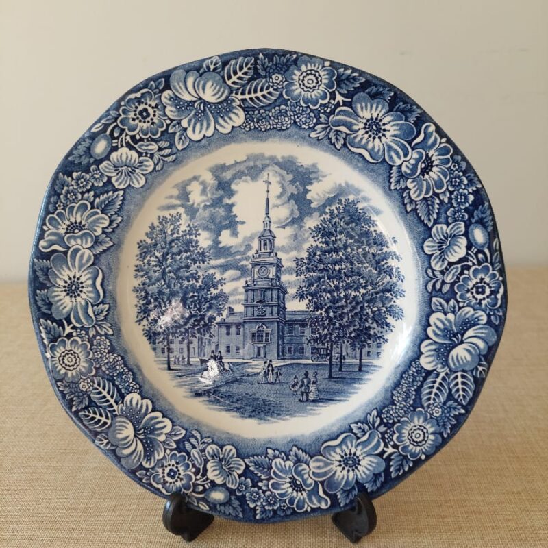 Vista frontal de un plato playo de loza inglesa Liberty Blue con el grabado central de la escena histórica de Independence Hall en azul cobalto y un ancho borde decorado con flores silvestres.