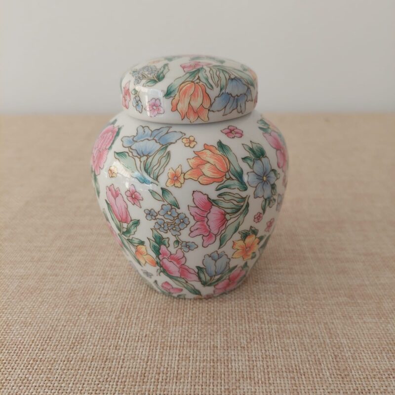 Vista frontal de un potiche miniatura de porcelana blanca con tapa, decorado con ramilletes de flores, tulipanes y pensamientos en tonos pastel rosa, naranjo y azul.