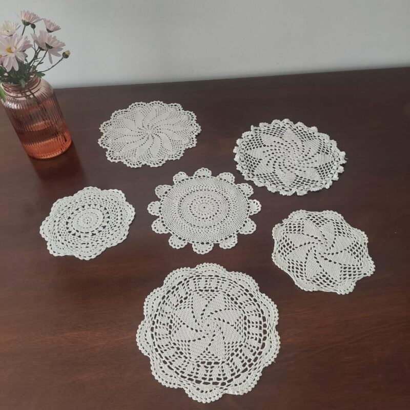 Vista de detalle de 6 tapetes circulares de encaje blanco tejidos a mano a crochet con diferentes patrones geométricos calados, presentados en hilera sobre una superficie de madera oscura.