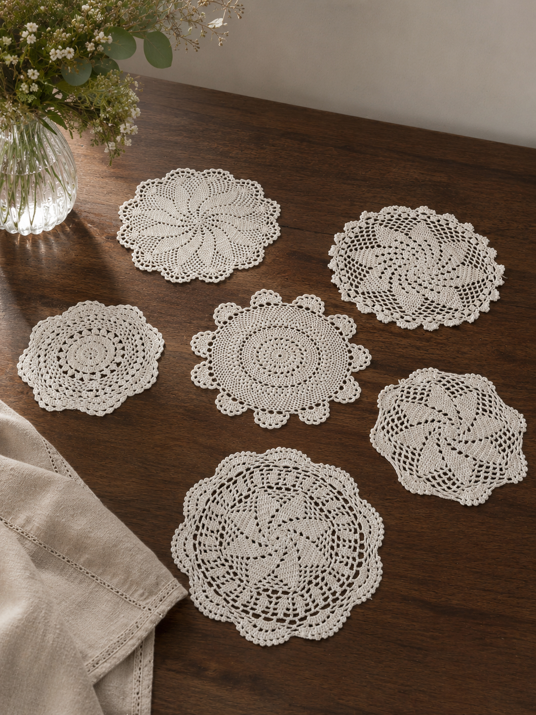 Vista de detalle de 6 tapetes circulares de encaje blanco tejidos a mano a crochet con diferentes patrones geométricos calados, presentados en hilera sobre una superficie de madera oscura.