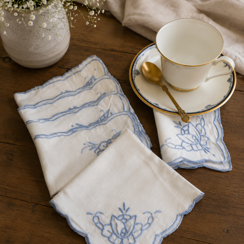 Vista superior de un set de servilletas blancas vintage de té con bordados y calados de rosas en azul pastel; una servilleta se dobla decorativamente debajo de una taza y platillo de porcelana con bordes dorados, mientras las otras cuatro descansan apiladas a un costado.