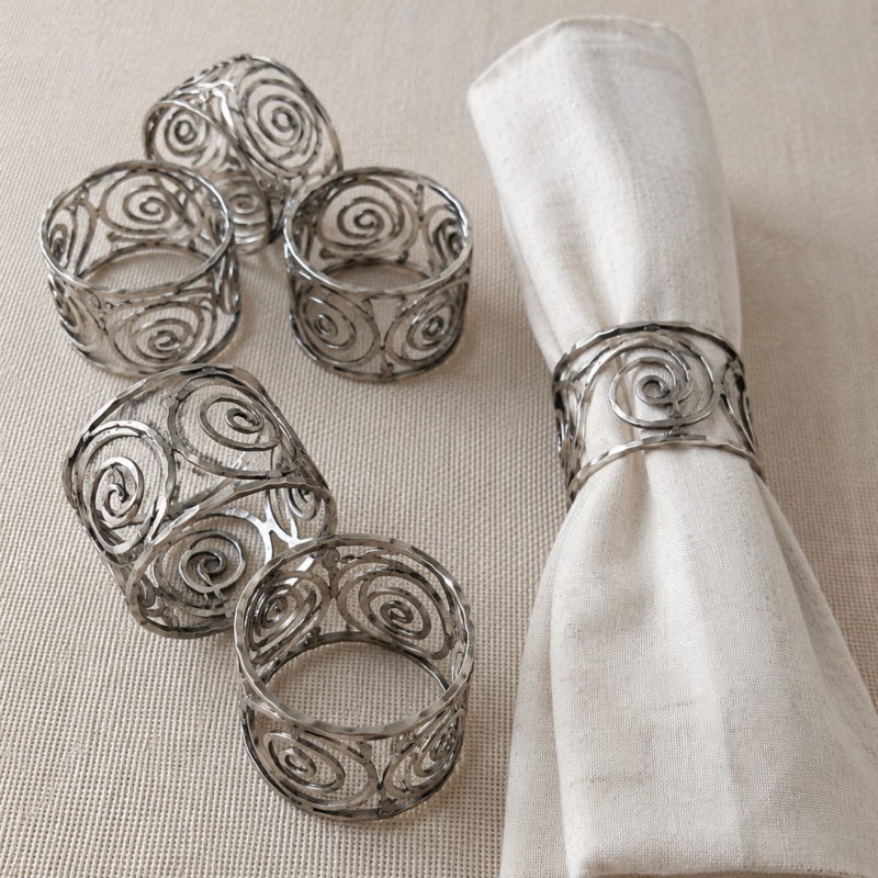 Set de seis anillos para servilleta de metal calado color plateado con diseño de espirales decorativas, presentados junto a una servilleta de lino en tonos naturales.