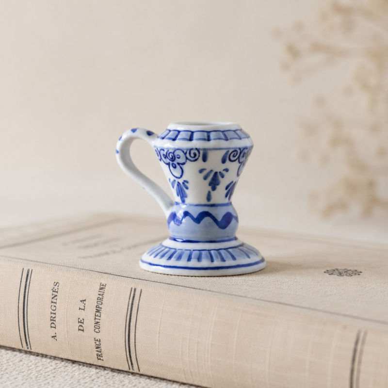 Primer plano de un pequeño candelabro palmatoria vintage de cerámica blanca holandesa con asa. Decorado con patrones botánicos y geométricos pintados a mano en color azul cobalto, sobre una superficie lisa y brillante apoyada en un fondo neutro.