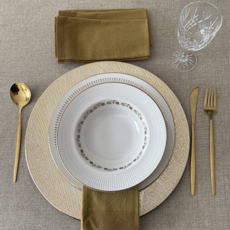 Vista cenital de un montaje de mesa rústico donde una servilleta de algodón color mostaza nueva cuelga elegantemente debajo de un plato hondo blanco con filigrana dorada, dispuesto sobre un plato base dorado; arriba se aprecian las otras tres servilletas dobladas, una copa de cristal y cubiertos de color dorado mate.