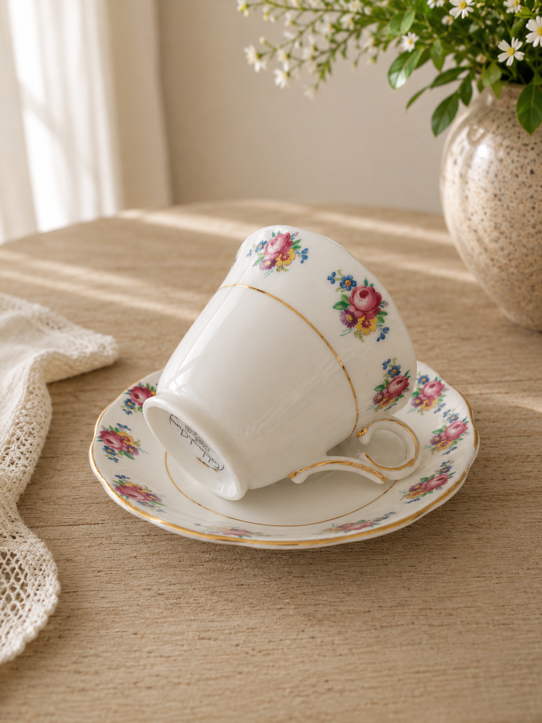 Vista de tres cuartos de una taza de té Colclough blanca recostada de lado sobre su plato a juego. Muestra guirnaldas de rosas fucsias y amarillas en el borde exterior, un asa contorneada dorada y el sello original negro en la base de la taza, sobre un paño beige.