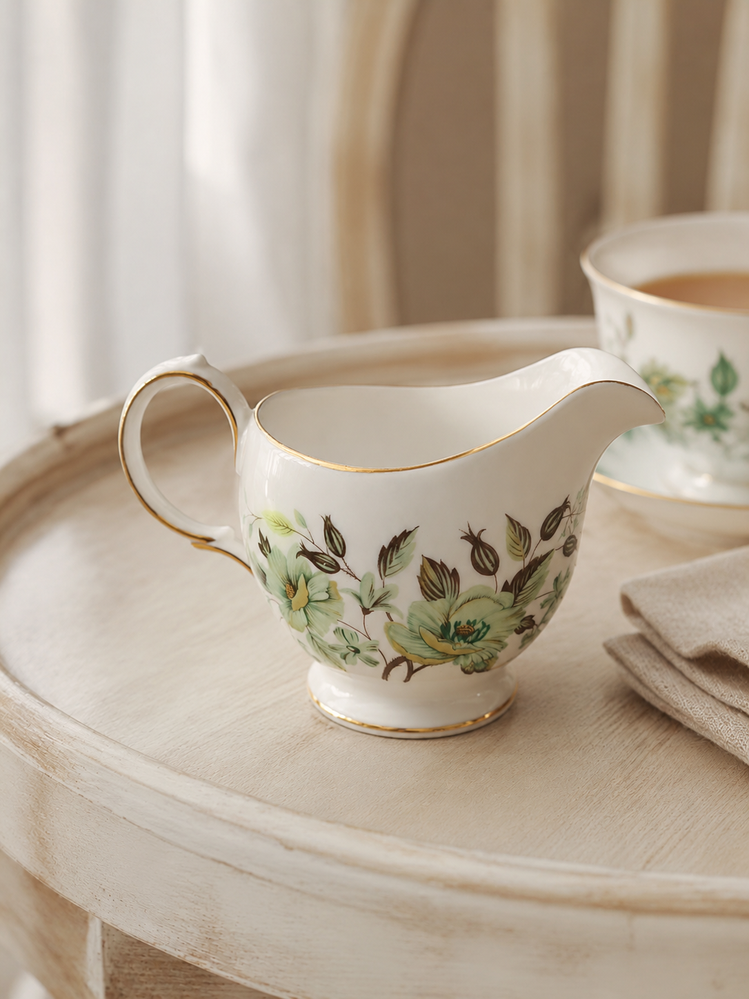 Vista lateral de una jarrita lechera de porcelana fina inglesa de la marca Colclough modelo Sedgley dispuesto sobre un paño beige. La pieza es blanca con un patrón de grandes flores verde menta y hojas café pintadas al frente, un pico vertedor pronunciado, un asa curva contorneada y finos filos dorados brillantes en sus bordes ondulados.