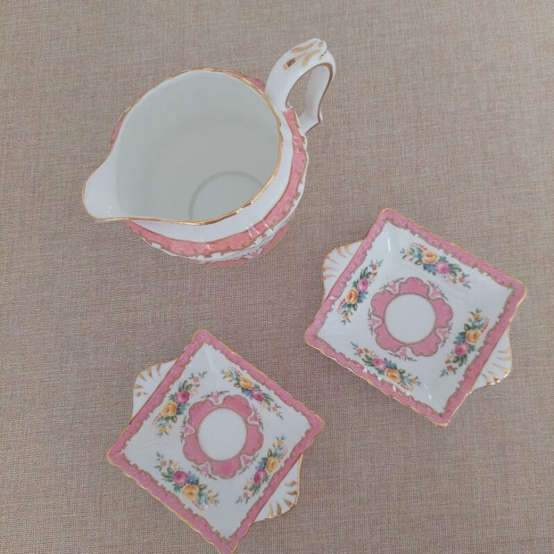 Vista frontal de tres cuartos de una jarrita cremera de porcelana fina de hueso de la marca inglesa Crown Staffordshire. Luce un fondo blanco níveo con una franja rosa decorada con filigranas, una vistosa guirnalda central con flores amarillas, rosadas y azules, un asa contorneada con acabados en oro y un borde ondulado impecable, posada sobre un fondo neutro beige.