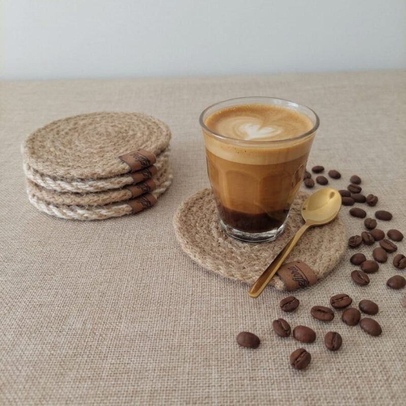 Vista de estilo de vida sobre una mesa de arpillera beige. Se muestra un posavasos redondo de yute trenzado sosteniendo un vaso de vidrio con un café cortado y arte latte de corazón, acompañado de una cuchara dorada y granos de café esparcidos. Al fondo a la izquierda, se aprecia una pila con los otros cinco posavasos del set mostrando sus etiquetas de ecocuero.