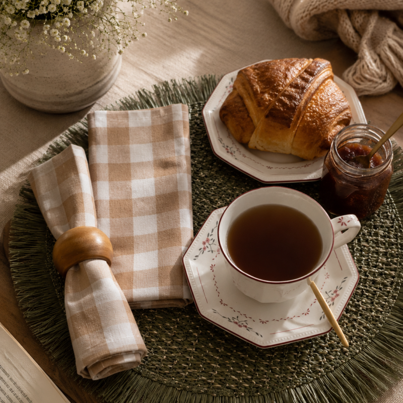 Vista cenital de un montaje de mesa para el té donde una servilleta nueva a cuadros Vichy camel y blancos se luce enrollada con un servilletero de madera cilíndrico sobre un individual redondo de color verde con flecos, al costado de una taza de porcelana con platillo octogonal decorada con flores rosadas.