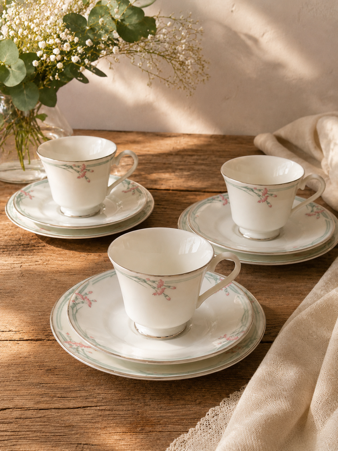 Composición en perspectiva de tres tríos de té de porcelana fina blanca Royal Doulton modelo Carnation. En primer plano se aprecia un trío completo con su taza, platillo y plato plano con guirnaldas suaves grisáceas y flores rosadas, mientras los otros dos tríos complementarios se observan de fondo simétricamente ordenados sobre una superficie textil beige.