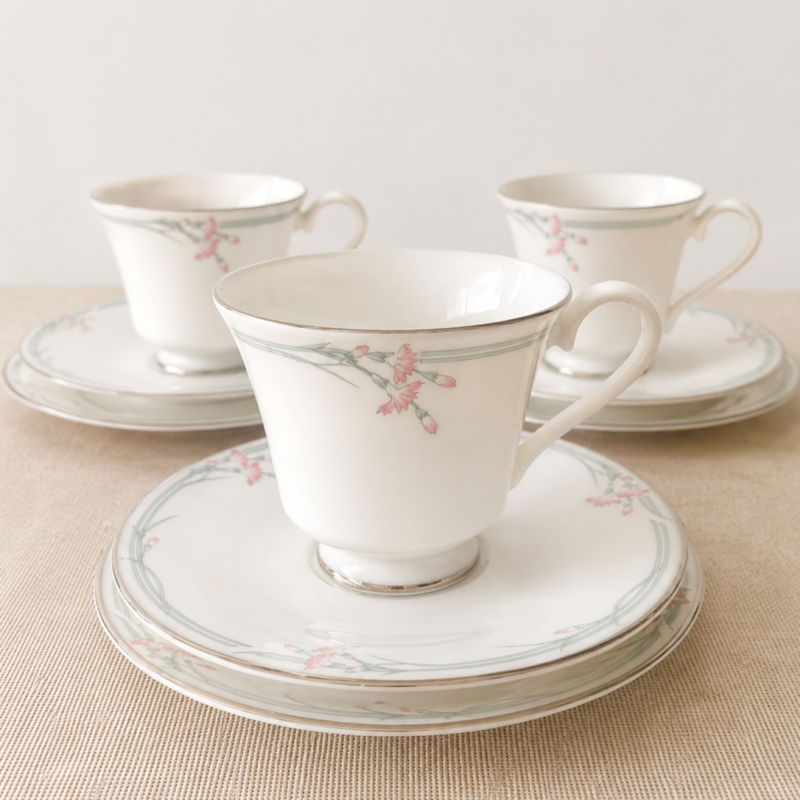 Composición en perspectiva de tres tríos de té de porcelana fina blanca Royal Doulton modelo Carnation. En primer plano se aprecia un trío completo con su taza, platillo y plato plano con guirnaldas suaves grisáceas y flores rosadas, mientras los otros dos tríos complementarios se observan de fondo simétricamente ordenados sobre una superficie textil beige.