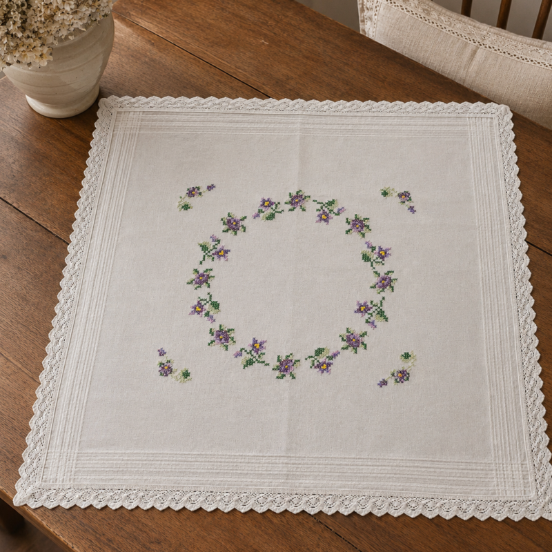 Vista cenital de una carpeta cuadrada blanca de estilo europeo nueva extendida, decorada con una gran corona central ovalada de flores silvestres en tonos violeta y lila bordada en punto cruz, bordes con alforzas y puntilla festonada tejida a crochet.