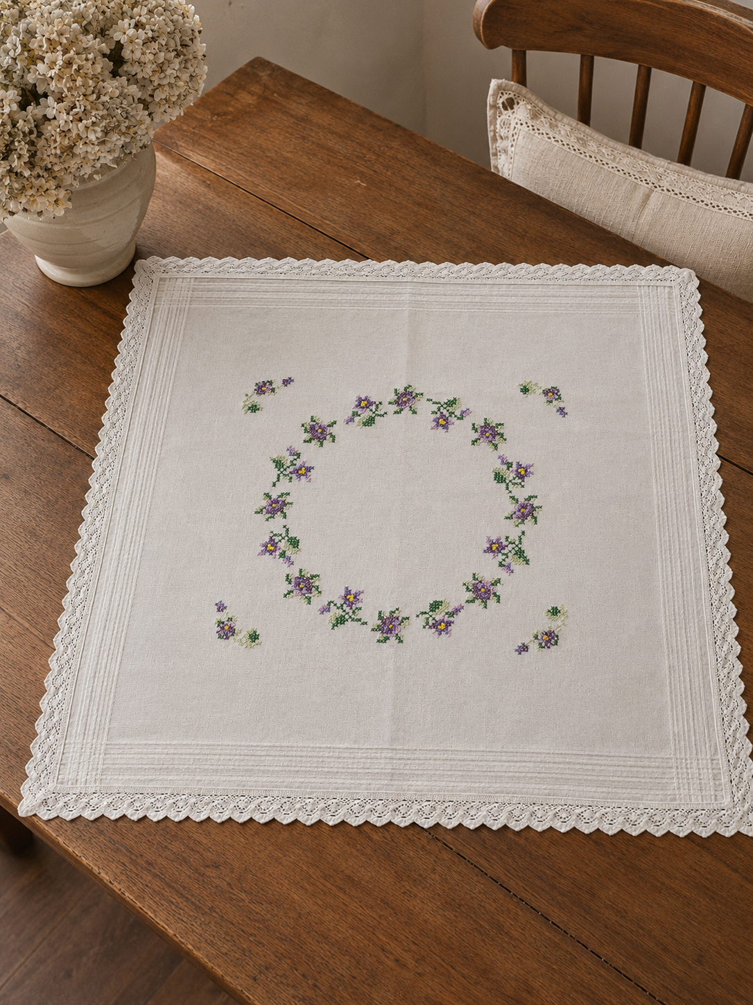 Vista cenital de una carpeta cuadrada blanca de estilo europeo nueva extendida, decorada con una gran corona central ovalada de flores silvestres en tonos violeta y lila bordada en punto cruz, bordes con alforzas y puntilla festonada tejida a crochet.