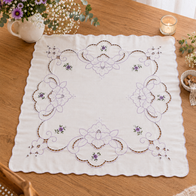 Vista cenital completa de una carpeta de mesa cuadrada de algodón blanco dispuesta sobre una mesa de madera clara. Destaca una gran corona cuadrangular calada estilo Richelieu delineada en hilo lila con pequeños ramilletes de flores violetas bordadas, ambientada con un florero blanco con flores moradas y una vela encendida.