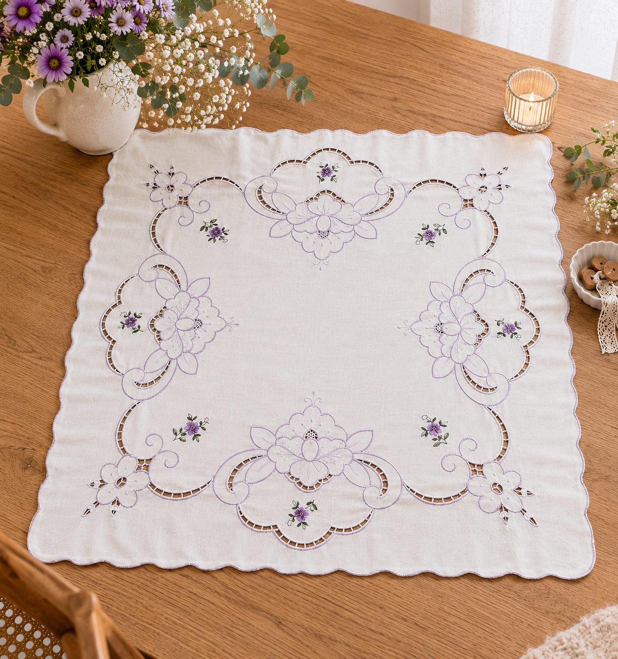 Vista cenital completa de una carpeta de mesa cuadrada de algodón blanco dispuesta sobre una mesa de madera clara. Destaca una gran corona cuadrangular calada estilo Richelieu delineada en hilo lila con pequeños ramilletes de flores violetas bordadas, ambientada con un florero blanco con flores moradas y una vela encendida.