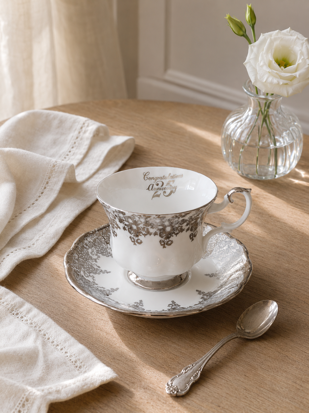 Vista frontal de un elegante dúo de té de porcelana fina inglesa Royal Albert modelo 25th Anniversary sobre un paño beige. Muestra una taza y plato blancos con un relieve ondulado, decorados con una ancha guarda perimetral plateada en filigrana, asa y base de platino brillante, y la frase manuscrita "Congratulations 25th Anniversary" en el interior de la taza.